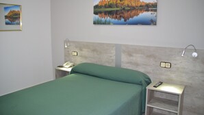 Una habitación de hotel con una cama, una mesita de noche con un teléfono, una lámpara y un cuadro de un lago con árboles otoñales.