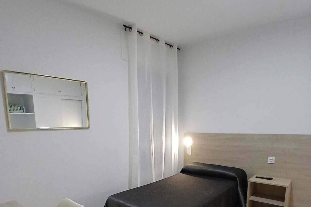Une chambre d’hôtel avec un lit, une chaise, une table de chevet, un miroir et une fenêtre avec des rideaux.