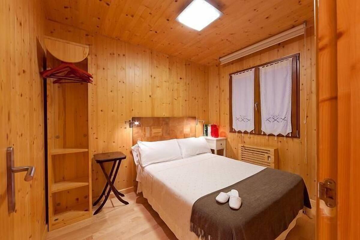 A wooden room with a bed, a small table, a shelf, and a window with curtains.