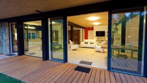 A modern house with a wooden deck, large glass doors leading to a living area with a sofa and chairs, and a kitchen visible through the glass. - Las casas de Ea Astei (Ea)