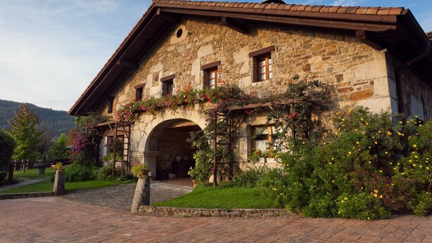 A stone building with a tiled roof, an archway entrance, and a garden with flowers and shrubs. - Las casas de Ea Astei (Ea)