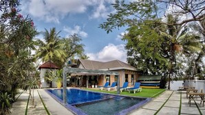 A swimming pool with blue tiles, surrounded by a concrete deck with green grass patches. There are lounge chairs and a covered area with a tiled roof. - Salty River Resort (Kampot)