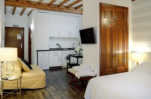 A hotel room with a bed, a bedside table with a lamp, a sofa, a wooden wardrobe, a television mounted on the wall, and a kitchenette with a washing machine and a sink.