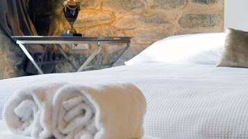 A neatly made bed with a rolled white towel and a folded white towel, a bedside lamp, and a stone wall in the background.