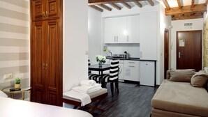 A compact hotel room with a wooden wardrobe, a dining area with a table and chairs, a kitchenette, and a sofa.