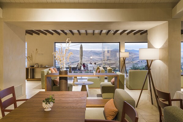 A spacious living area with a wooden dining table, comfortable seating, and large windows offering a view of the outdoors.