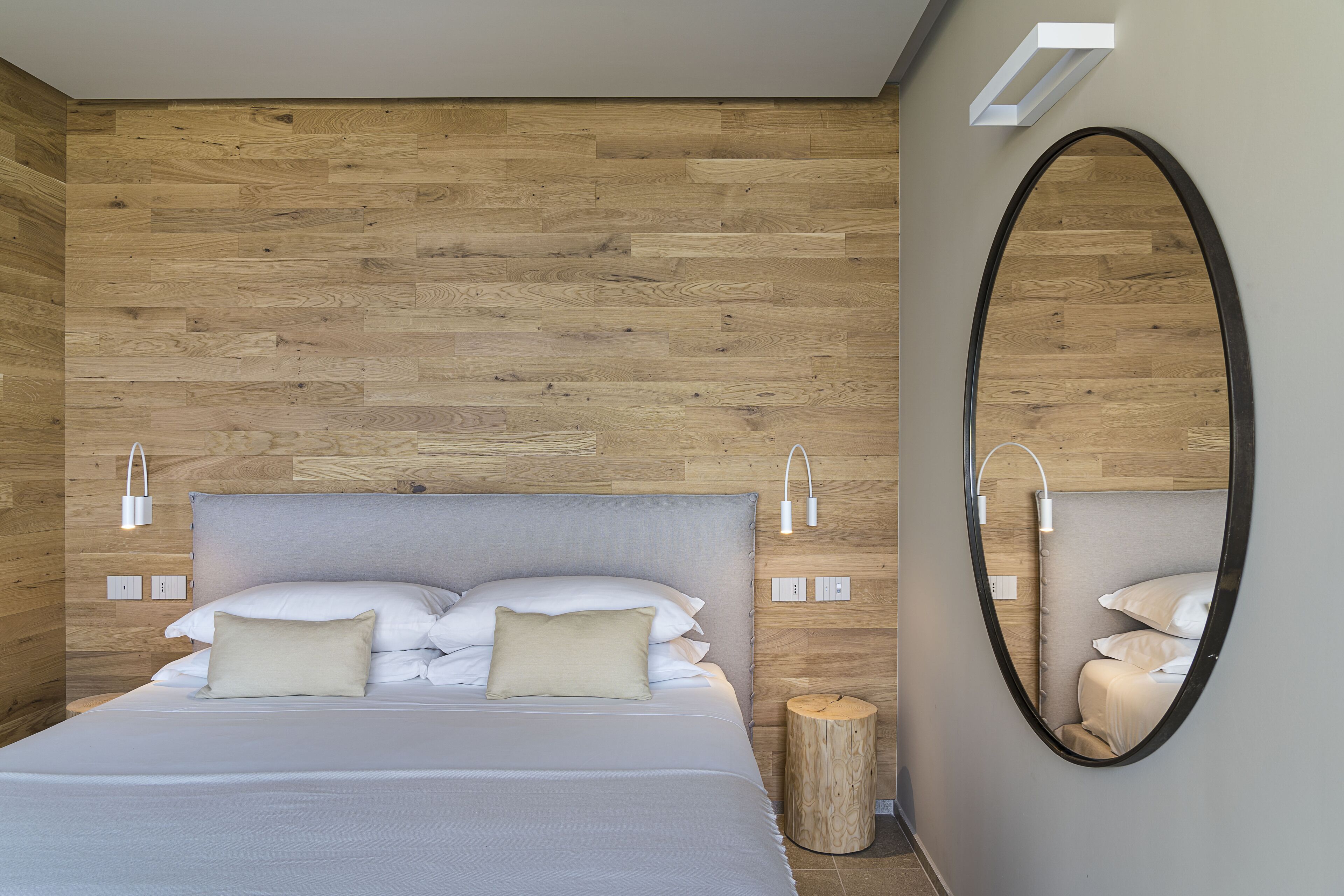 A modern bedroom with a wooden wall panel, a large bed with white bedding, a round mirror, and bedside lamps.