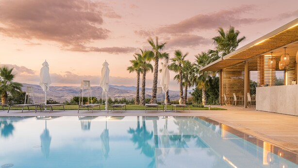 Zona de piscina con sillones de descanso, palmeras y vistas a montañas.