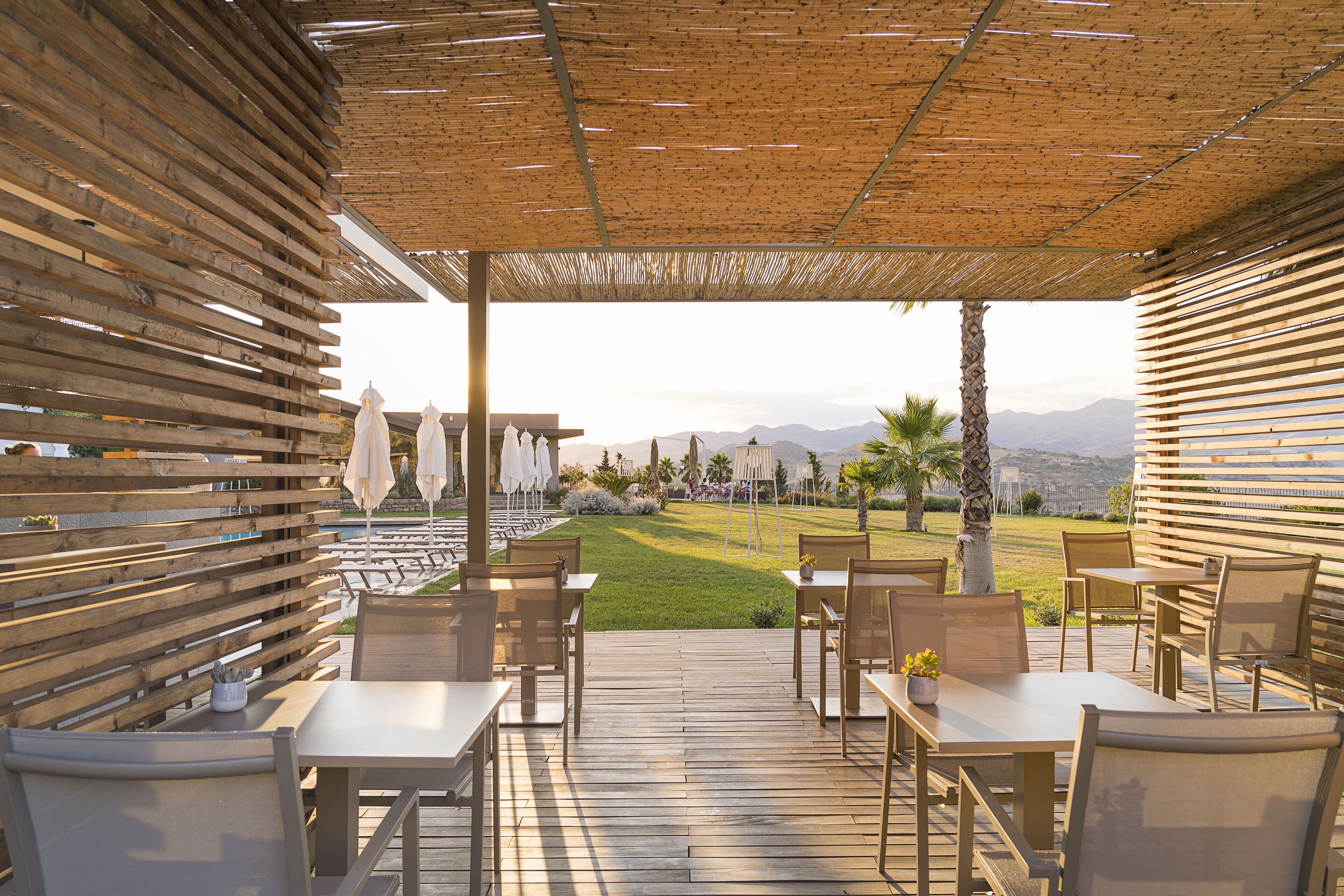 Un patio exterior con mobiliario de madera y sombrillas, con vistas a una zona verde con palmeras y montañas a lo lejos.