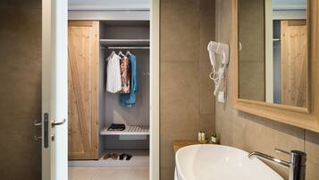 Un baño moderno con un lavamanos grande de color blanco, un secador de pelo montado en la pared y un armario de madera con ropa colgada.