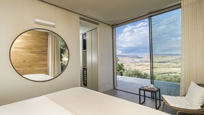 Un dormitorio con una cama grande, un espejo redondo, una silla y vistas a un paisaje a través de una puerta corrediza de vidrio.