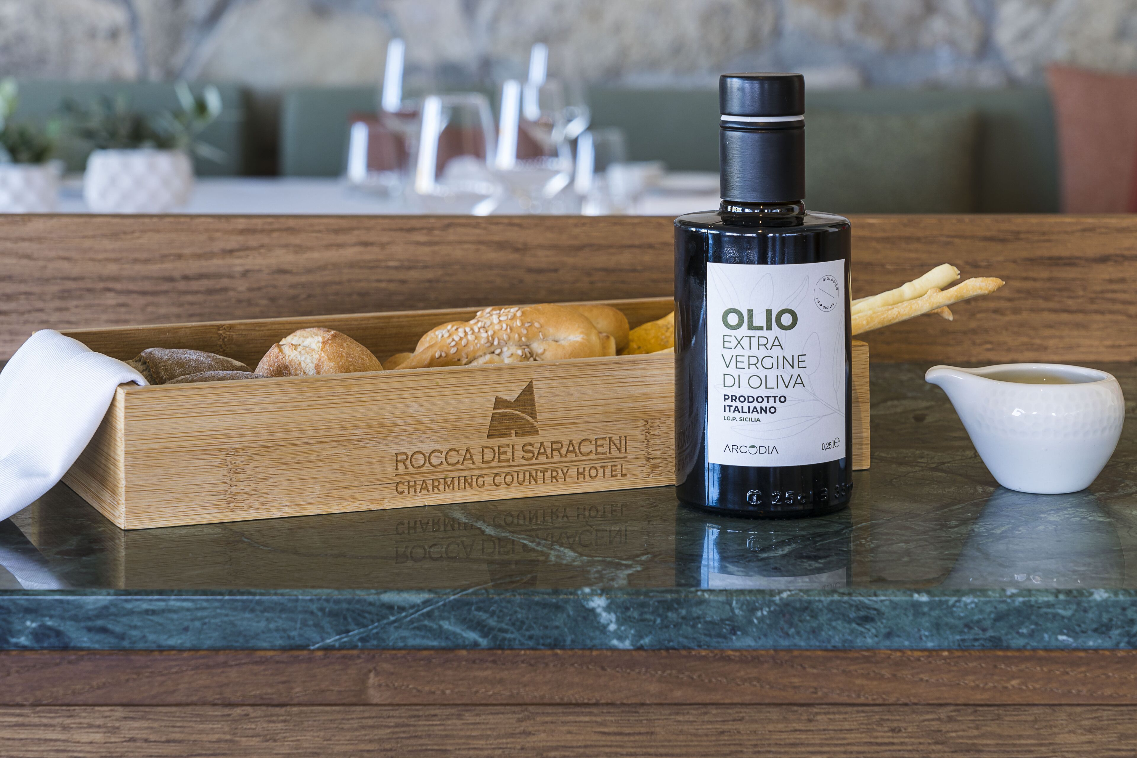 Una botella de aceite de oliva virgen extra, una caja de madera con pan y una pequeña jarra blanca sobre una encimera de mármol.
