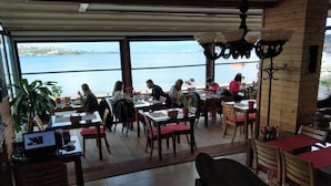Un restaurante con amplios ventanales que dan a una masa de agua, mesas y sillas de madera, y plantas en macetas.