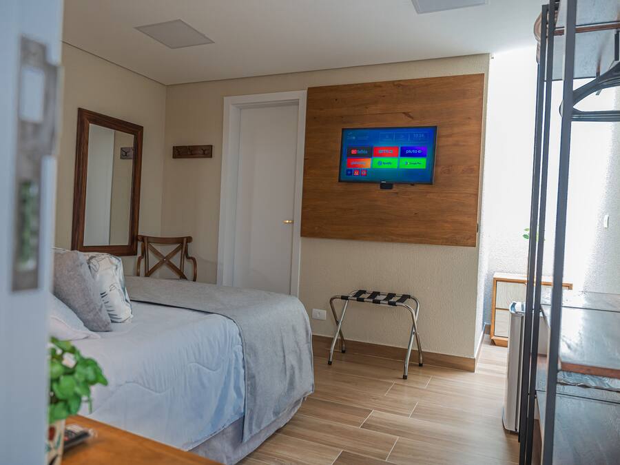 A hotel room with a bed, a TV mounted on the wall, a small stool, and a wooden table.