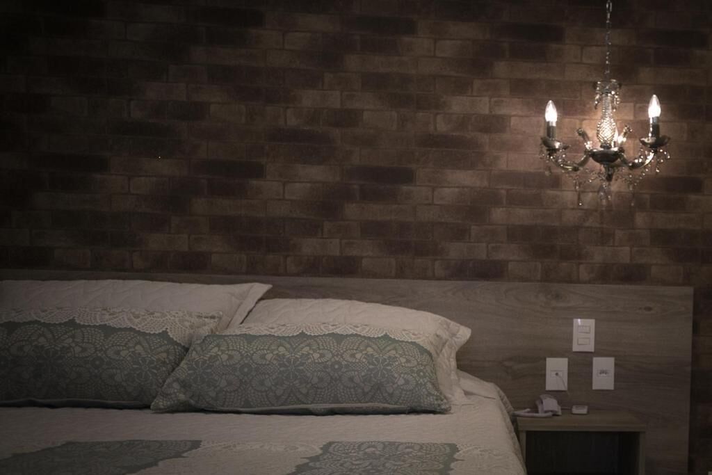 A bedroom with a brick wall, a chandelier, a bed with lace pillows, and a nightstand with a phone and a light switch.