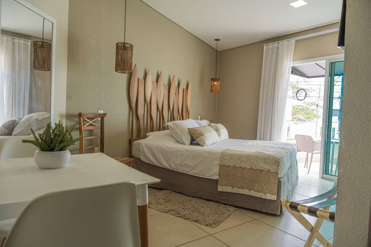 A bedroom with a bed, a bedside table, a chair, a potted plant, and a view of the outside.