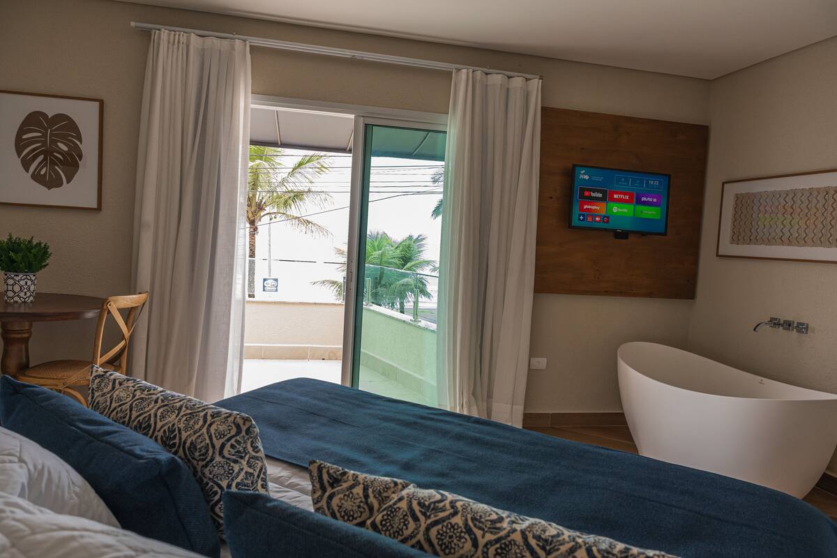 A modern hotel room with a large bed, a bathtub, a TV on the wall, and a balcony with curtains.