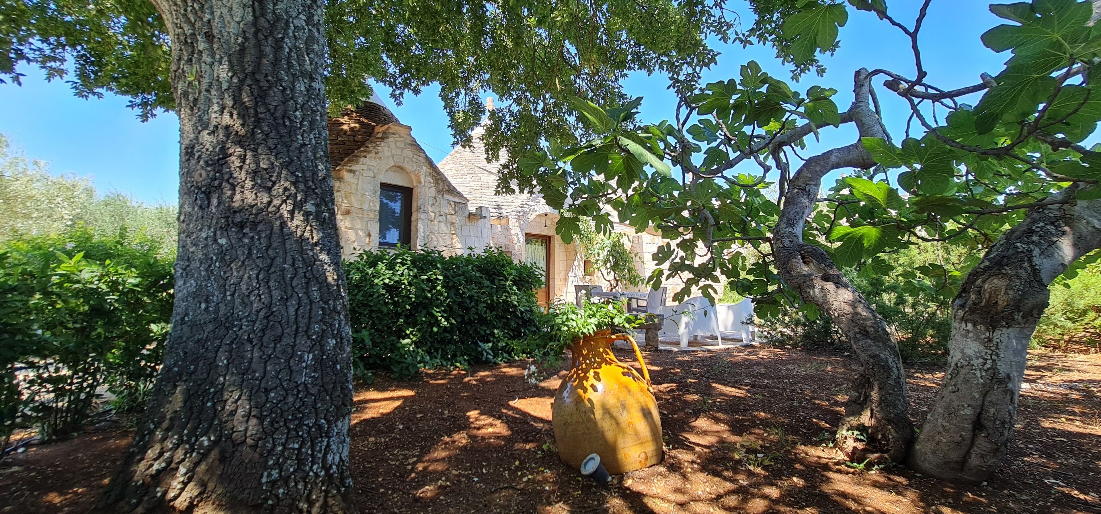 A stone building with a tiled roof, surrounded by trees and a garden area with a large pot and outdoor furniture.