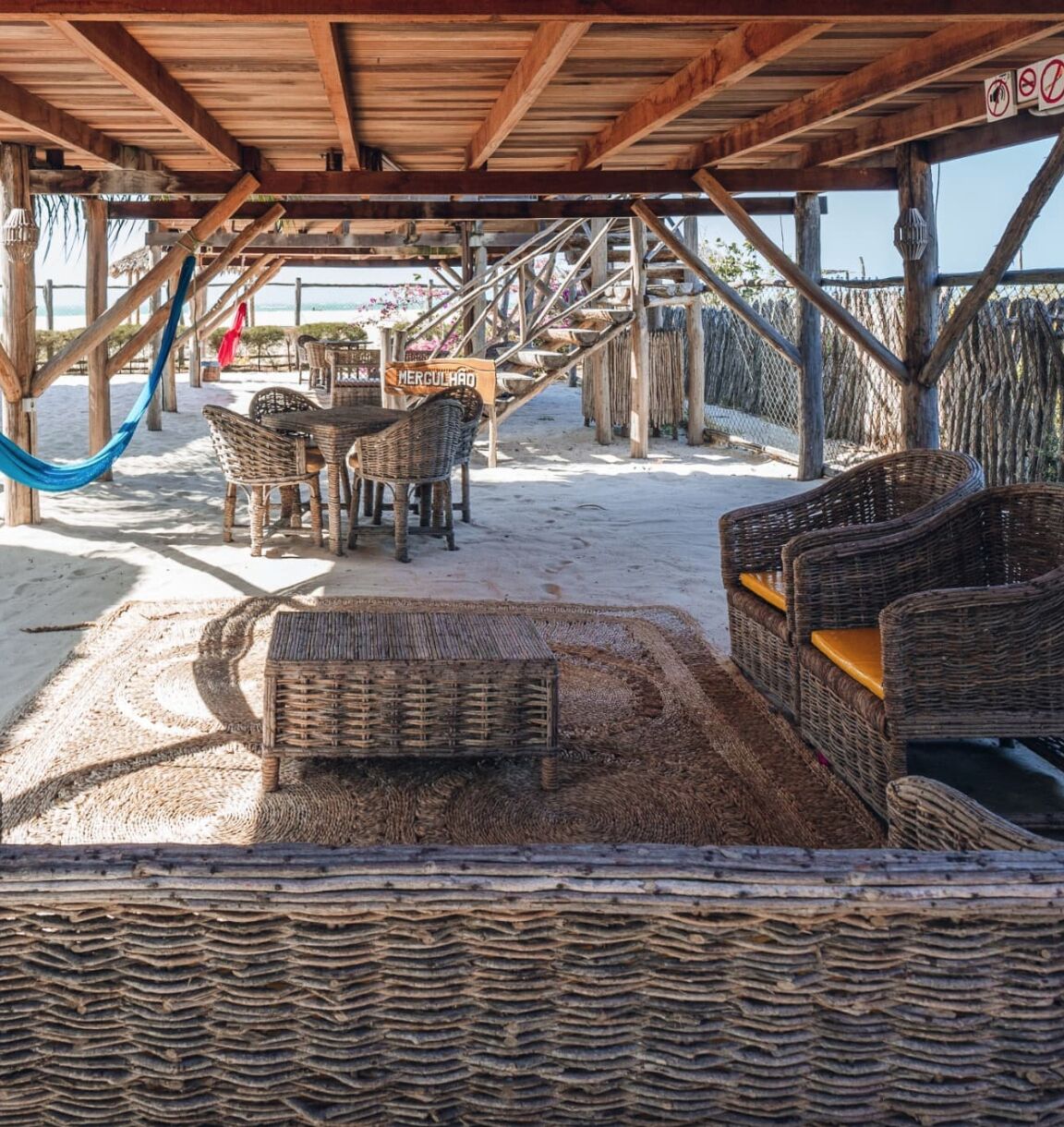 Una estructura de madera frente a la playa con mobiliario de mimbre, una hamaca y un letrero que dice "MERCULLO".