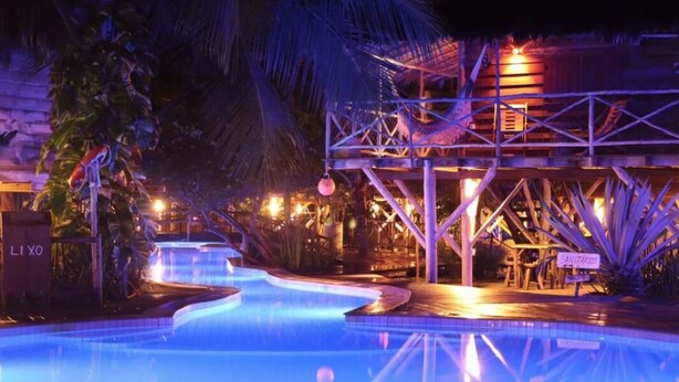 Zona de piscina nocturna con fuente, palmeras y una estructura con el letrero 'LI XO'.