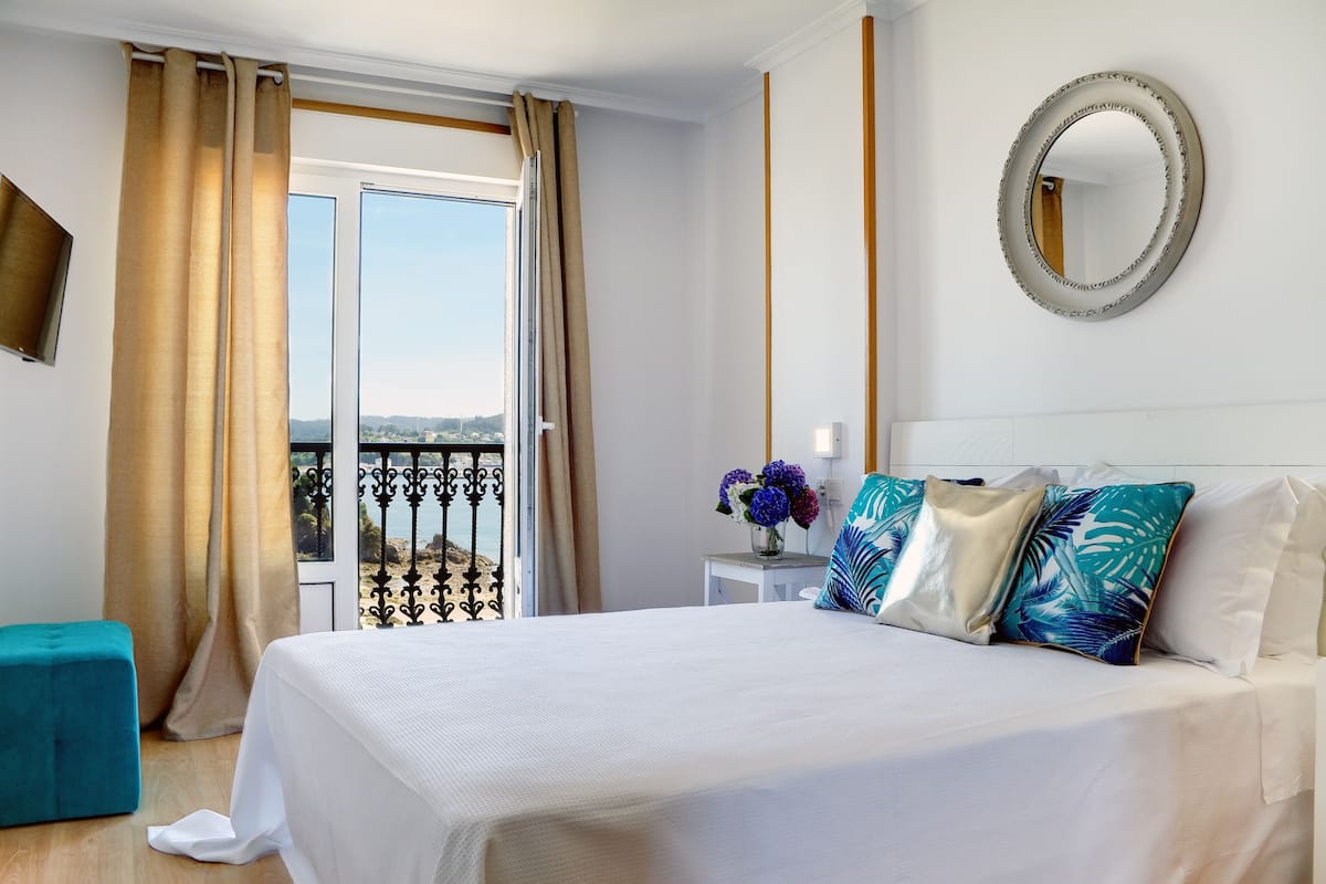 Quarto de hotel com cama, pequena mesa com um vaso de flores, varanda com cortinas e espelho fixado na parede.