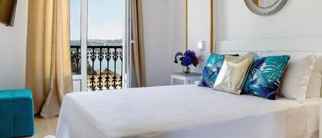 Habitación de hotel con cama, una mesita con un florero, un espejo en la pared y un balcón con cortinas.