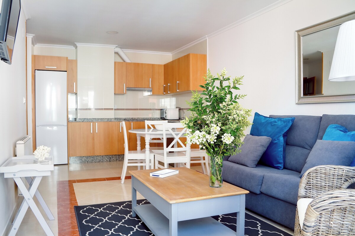 Una cocina moderna con armarios de madera, un refrigerador blanco y una zona de comedor con sillas blancas. Hay un sofá gris con cojines azules y una silla de mimbre. Una mesa de centro de madera con un jarrón de flores está en el centro.