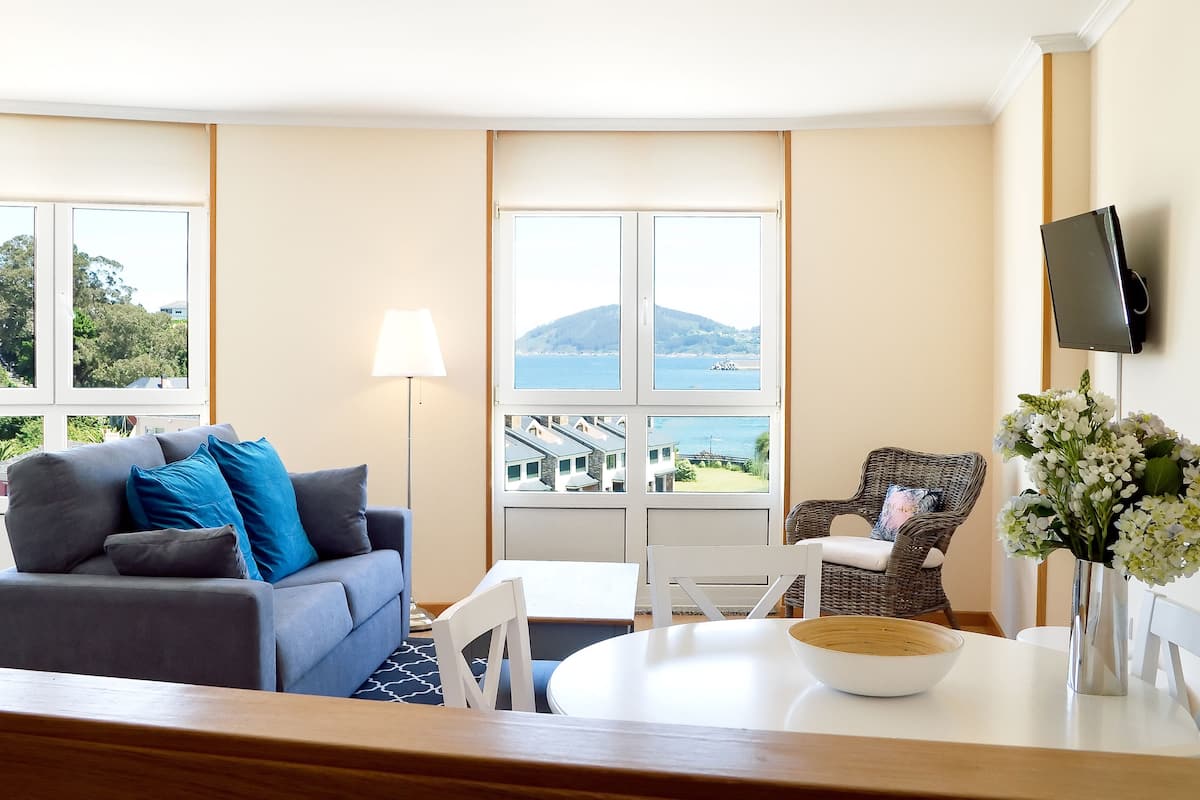 Quarto com sofá cinza, cadeira de vime, mesa de jantar com uma tigela e televisão na parede.