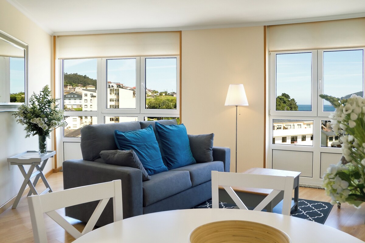 Una sala de estar moderna con un sofá gris, cojines azules, una mesa blanca y una lámpara de pie. Amplias ventanas ofrecen vistas a edificios y zonas verdes.