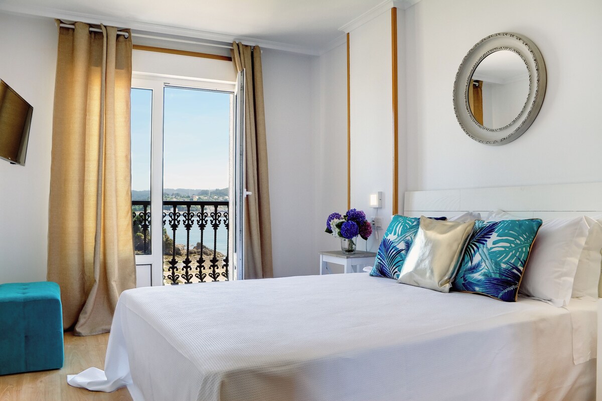 Habitación de hotel con cama, una mesita con un florero, un espejo en la pared y un balcón con cortinas.
