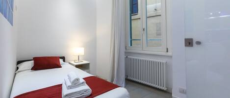 A hotel room with a bed, a nightstand, a lamp, a window with curtains, and a radiator.