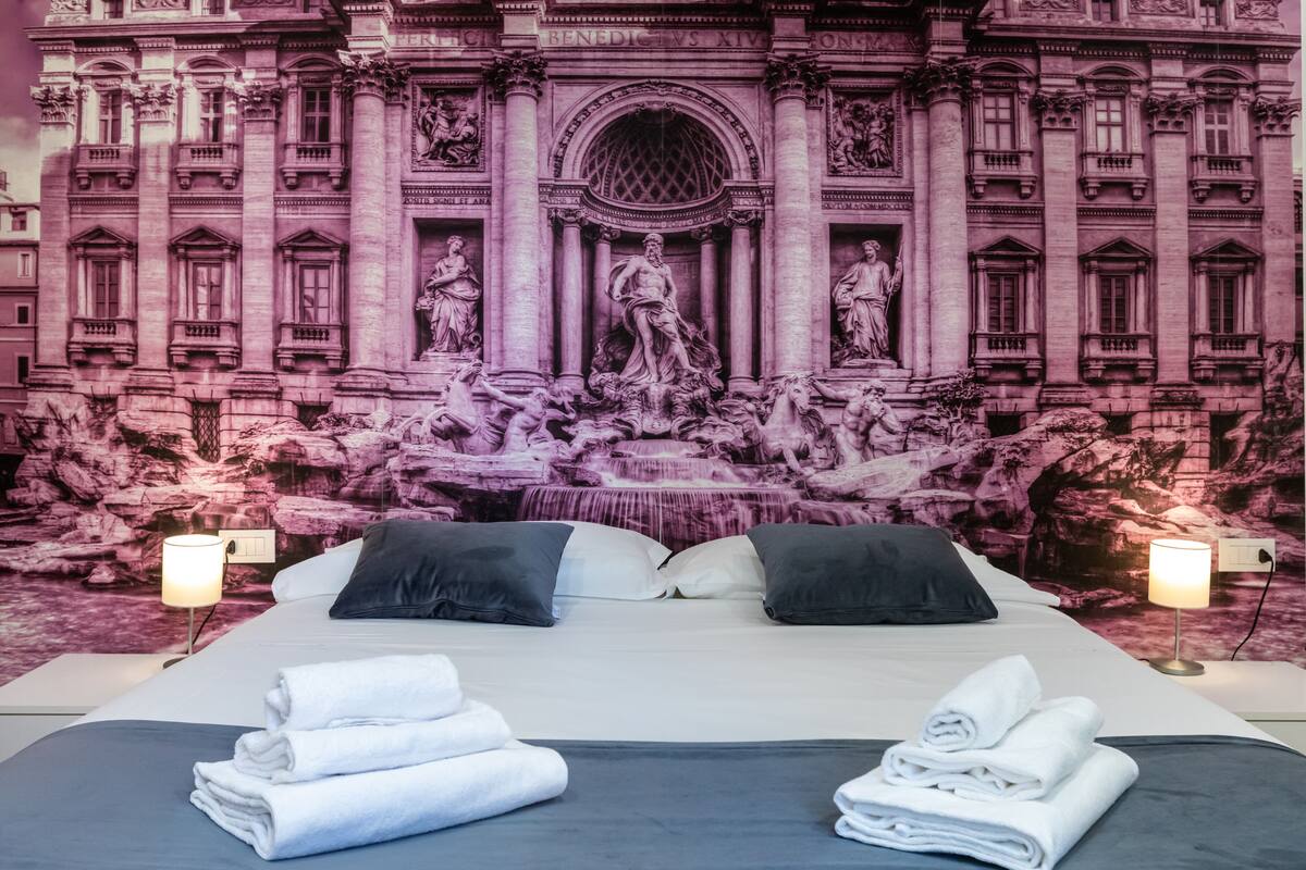 Habitación de hotel con una cama grande, dos lámparas de noche y un mural que representa una fuente y arquitectura clásica.