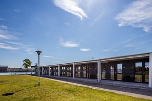 A row of modern, covered outdoor seating areas with numbered doors, a lamp post, and a view of a body of water in the background. - Smiltynes Jachtklubas (Klaipeda)