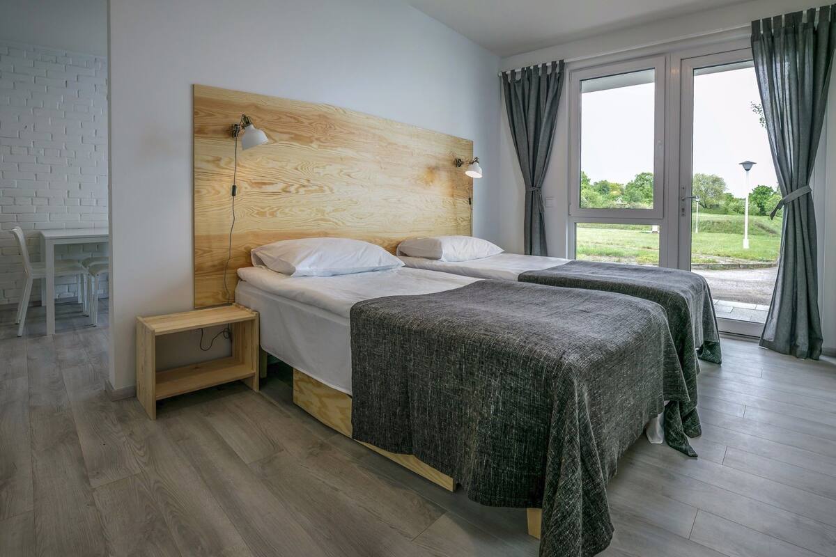 A modern bedroom with two beds, a wooden headboard, a bedside table, and a view of greenery through the window.