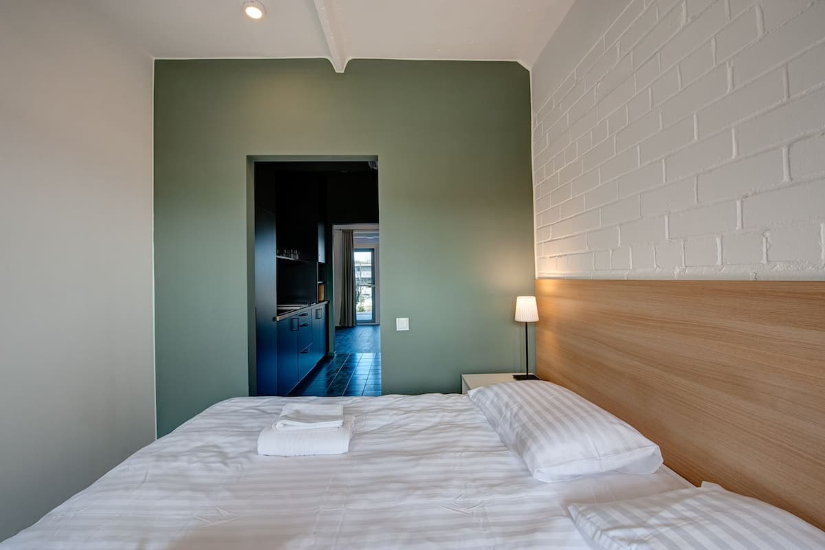 A hotel room with a bed, a nightstand, a lamp, and a view of a hallway leading to a balcony.