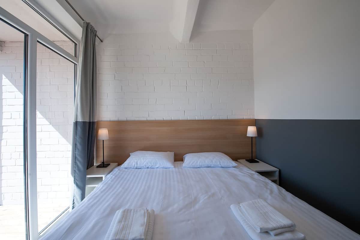 A hotel room with a bed, two bedside tables, a wooden headboard, and a large window with curtains.