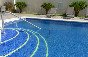 Una piscina azul cristalina con una escalera metálica, rodeada de sillones de descanso y palmeras.