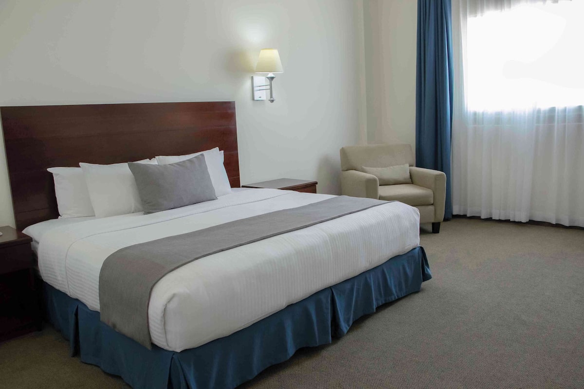 A hotel room with a large bed, a nightstand, a chair, and a lamp.