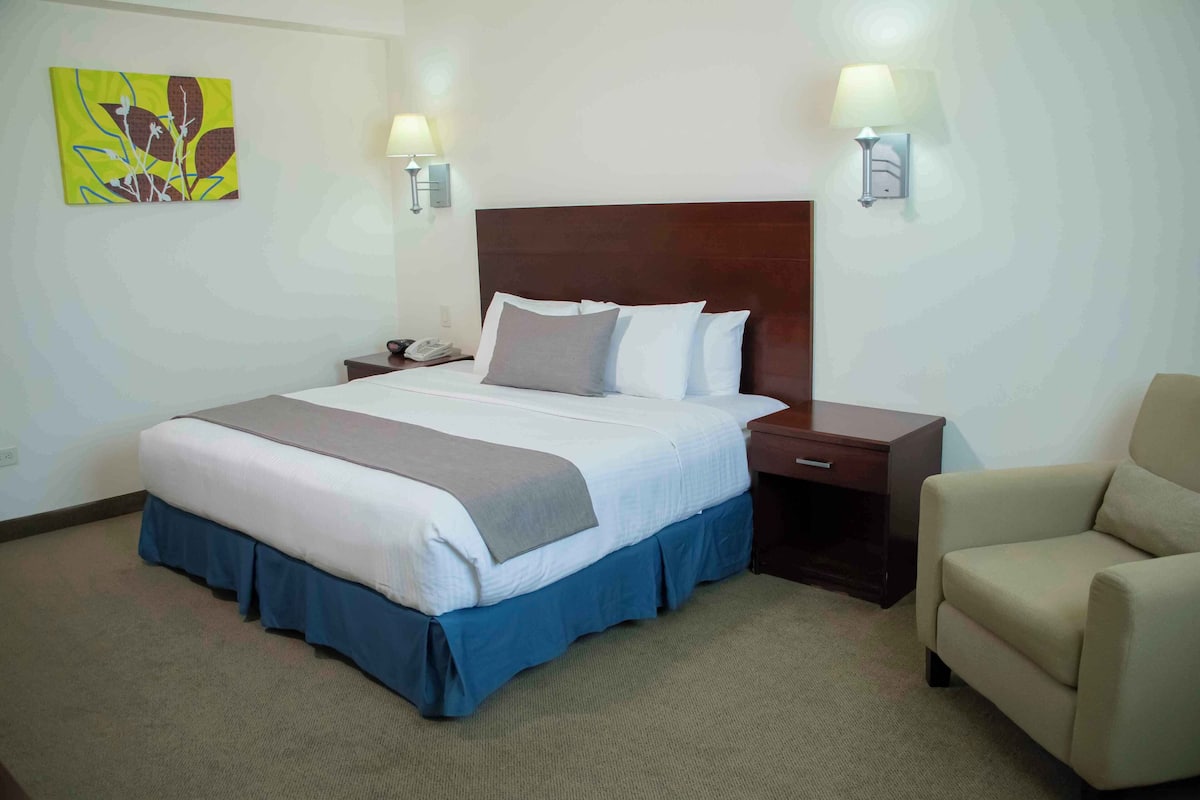 A hotel room with a large bed, bedside lamps, a desk, a chair, and a wall art piece.