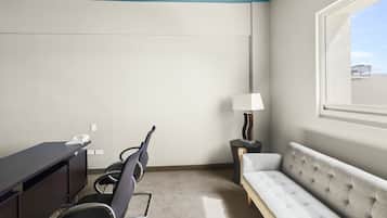 Habitación de hotel con un escritorio de madera oscura, una silla de oficina negra, un sofá blanco, una mesita auxiliar con una lámpara y un gran ventanal con vistas al cielo y un edificio.