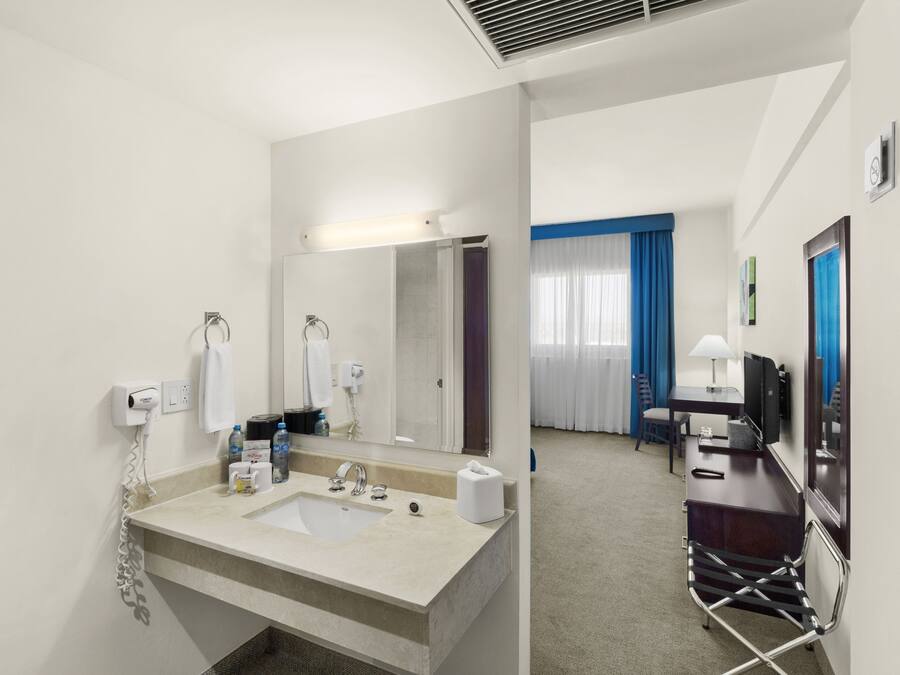 A hotel bathroom with a sink, mirror, towel dispenser, and a shower area.
