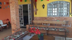 A room with a brick wall, a wooden bench, a floral sofa, a bench with red flowers, a window with a decorative frame, and a wall-mounted light fixture. - Albergue Piratas do Sol - Hostel (Recife)