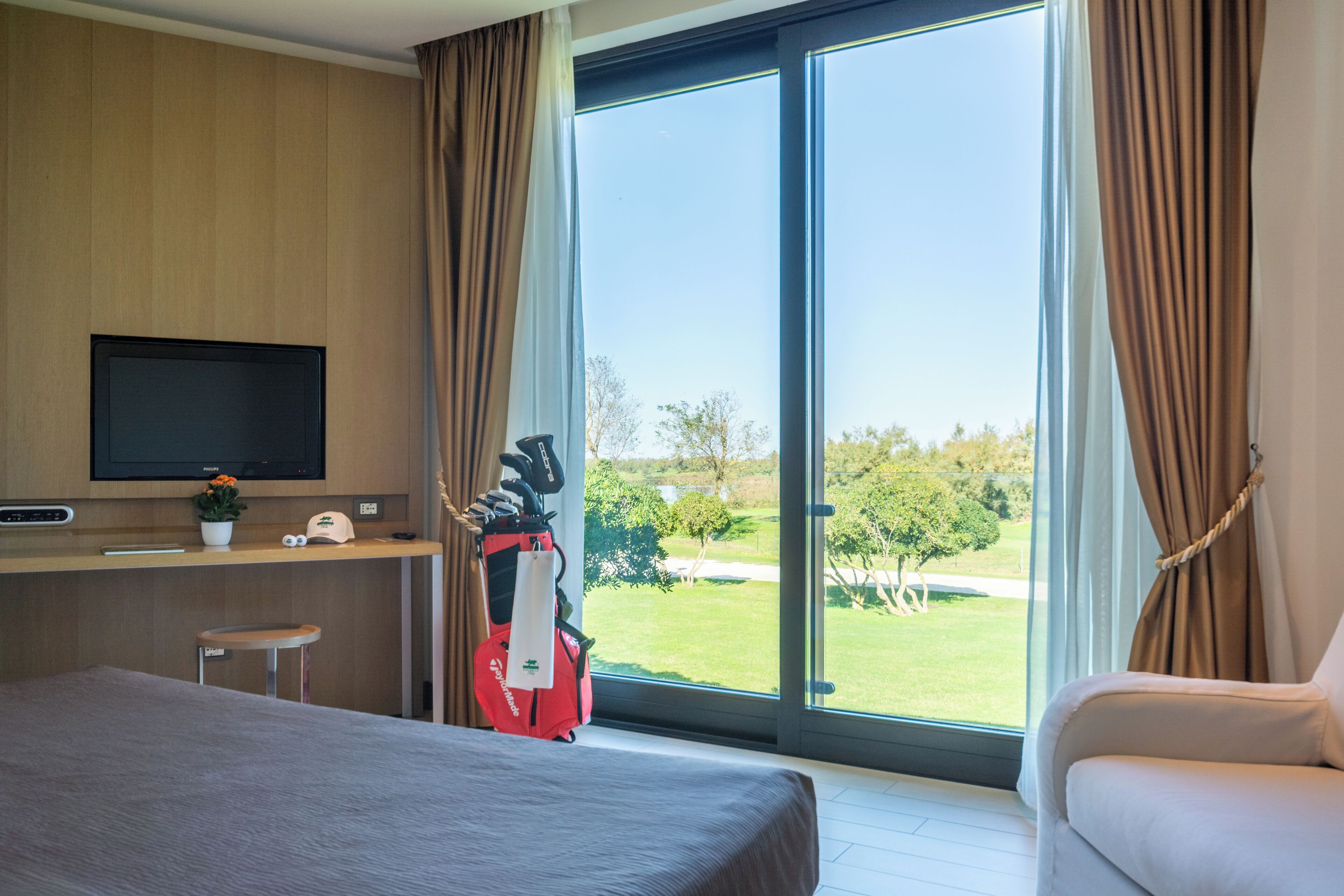 A modern hotel room with a bed, a desk with a TV, a red golf bag, and a view of a green landscape through the window.