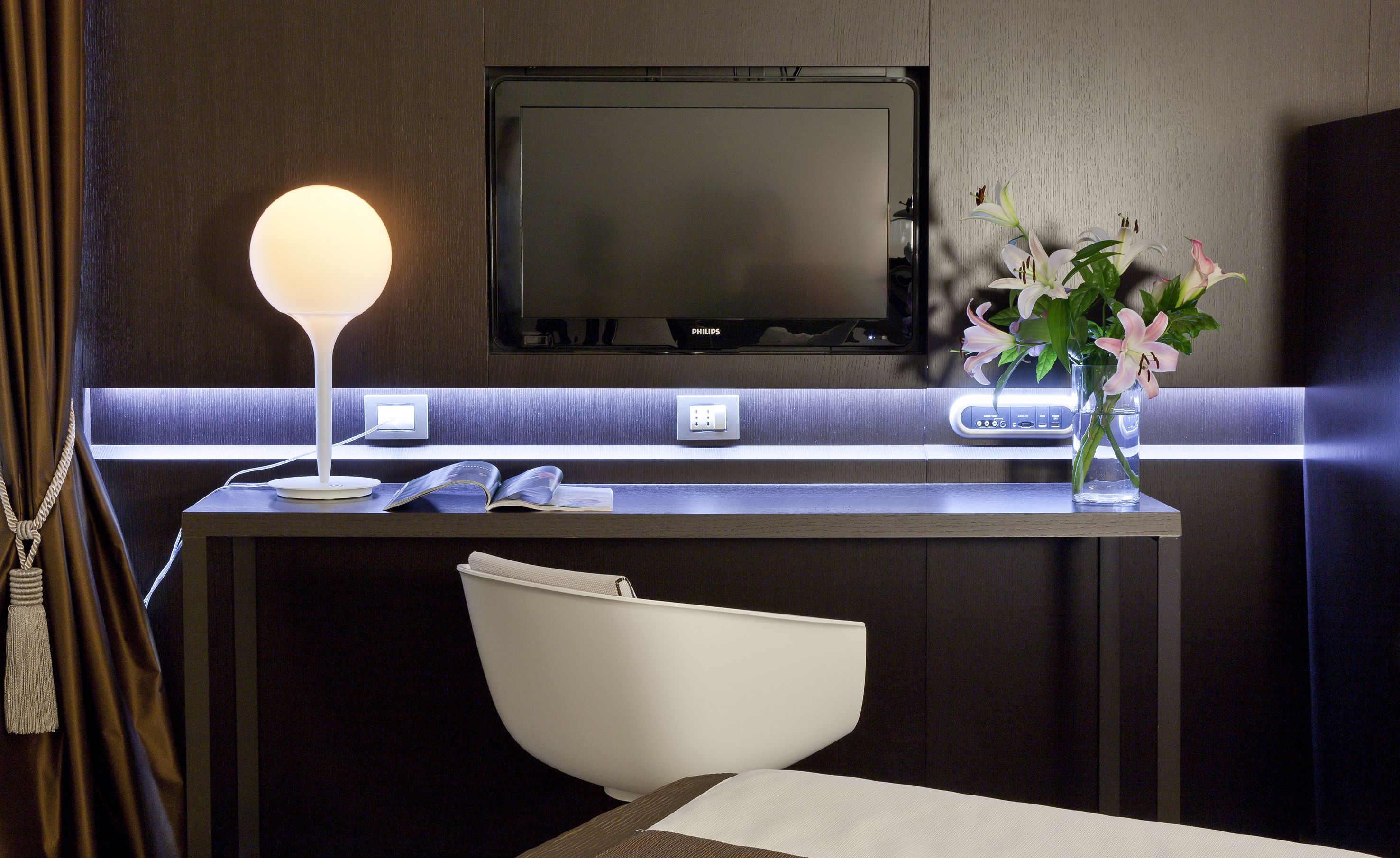 A hotel room with a desk, a lamp, a TV, a vase with flowers, and a chair.