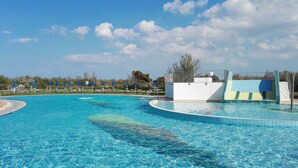 A clear blue swimming pool with a curved design, surrounded by blue umbrellas and a white building with a blue and yellow mural. - Isola di Albarella Golf Hotel (Rosolina)