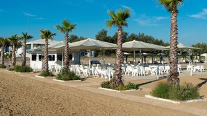 A beachside resort with outdoor seating areas, palm trees, and a clear blue sky. - Isola di Albarella Golf Hotel (Rosolina)