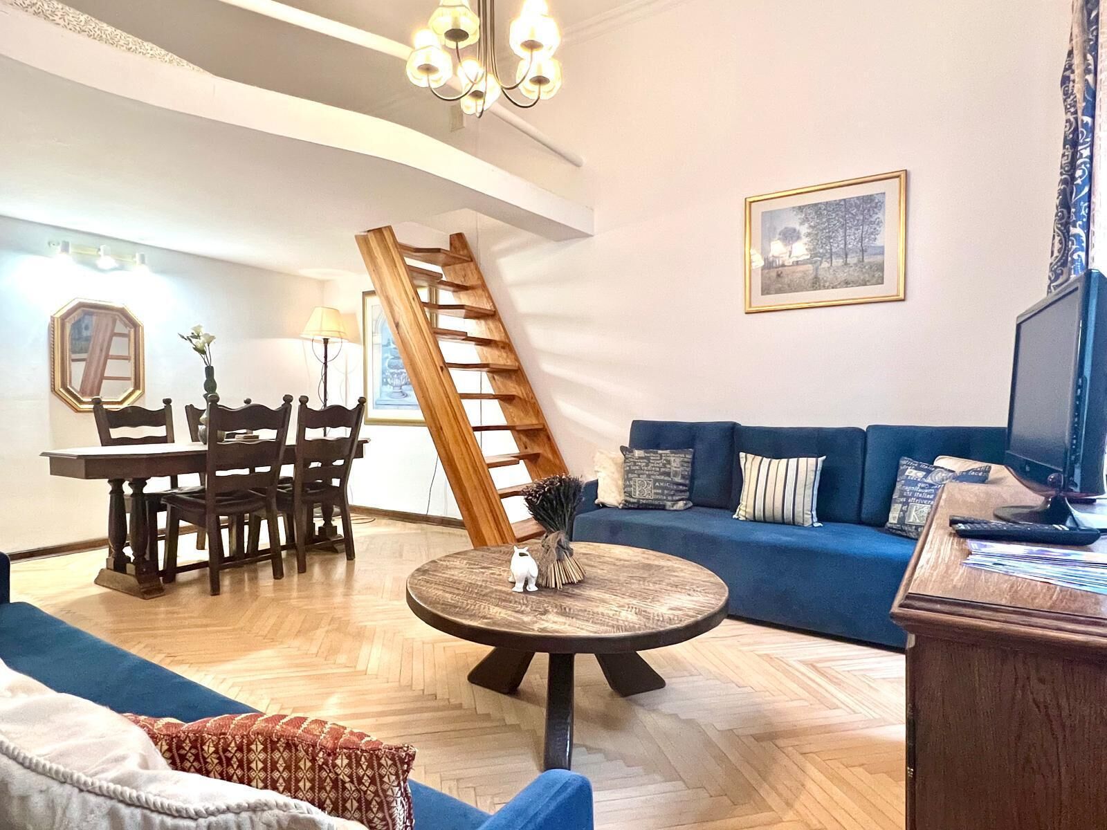 A living room with a blue sofa, a wooden dining table with chairs, a round coffee table, and a staircase.