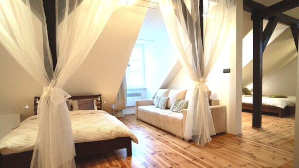 Un dormitorio con piso de madera, cama con dosel, sofá y ventana con luz natural.
