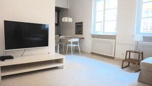 A modern living room with a flat-screen TV on a white stand, a dining area with a wooden table and white chairs, and a window with a view of the outside. - AAA STAY Castle Square Old Town (Warsaw)