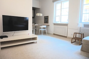 A modern living room with a flat-screen TV on a white stand, a dining area with a wooden table and white chairs, and a window with a view of the outside. - AAA STAY Castle Square Old Town (Warsaw)